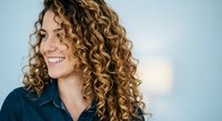 Young woman with voluminous curly blonde hair smiling in profile wearing denim shirt against soft blue background for lifestyle and beauty content.
