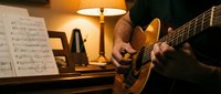 Musician playing acoustic guitar in cozy home studio with sheet music and warm lighting for practice session.