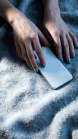 Woman's hands with blue nail polish holding a smartphone on a soft gray blanket, cozy lifestyle concept for social media.