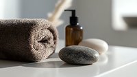 Minimalist spa composition with rolled brown towel, massage stone, and amber glass dispenser bottle on white surface in natural light. Wellness and relaxation concept.