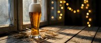 Golden beer glass with foam on rustic wooden table with wheat stalks and warm bokeh lights creating cozy pub atmosphere for brewing industry marketing.