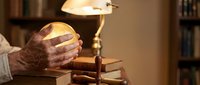 Senior adult hands holding glowing orb over stack of books under warm lamp light, symbolizing knowledge, wisdom, and learning in cozy home library setting.