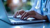 Healthcare professional typing on laptop computer, close-up of hands with stethoscope visible on blue medical uniform in dimly lit clinical environment.