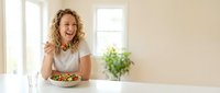 Happy young Caucasian woman eating healthy salad at kitchen table, smiling while enjoying nutritious meal in bright modern home interior.