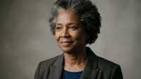 Professional African American businesswoman with natural gray afro hairstyle wearing dark blazer smiling confidently in corporate portrait for leadership concepts.