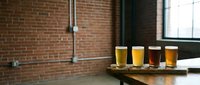 Craft beer flight with four glasses of different colored beers on wooden board in industrial brick brewery or pub setting for beverage, hospitality, and food service marketing.