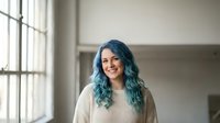 Young woman with vibrant blue hair smiling in bright modern interior space with large windows. Casual lifestyle portrait for wellness and self-expression concepts.
