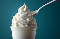 Swirled soft serve vanilla ice cream in white paper cup with plastic spoon against turquoise background, close-up view showing creamy texture and peaks.