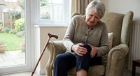Senior Caucasian woman sitting in comfortable armchair using smartphone at home with walking cane nearby. Elderly lifestyle and modern technology concept.