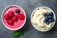 Fresh raspberry and blueberry smoothie bowls topped with whole berries and whipped cream on dark background with mint leaf garnish.