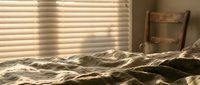 Cozy unmade bed with rumpled sheets and warm sunlight streaming through window blinds creating peaceful morning atmosphere in bedroom interior.