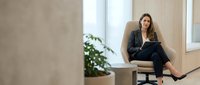 Young professional businesswoman sitting on desk in modern office space with laptop computer working remotely in bright natural light environment.