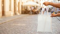 Person with medium skin tone holding white canvas tote bag on cobblestone street with blurred urban background, suitable for retail, shopping, and lifestyle marketing.