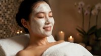 Young Asian woman enjoying relaxing spa treatment with white facial mask in candlelit wellness center for skincare and beauty routine.