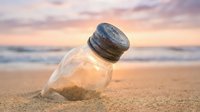 Glass bottle buried in golden beach sand at sunset with soft pink and purple sky creating romantic coastal atmosphere for environmental concepts.