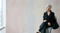 Senior woman with short gray hair and fair complexion wearing black blazer and pants, seated by window in modern office setting, looking away thoughtfully for corporate business concepts.