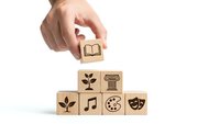 Hand placing wooden cube with book icon on stack of blocks featuring education and arts symbols on white background for learning concepts.