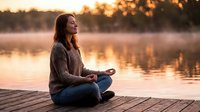 Young woman meditating in lotus position on wooden dock at sunset by lake. Peaceful mindfulness practice during golden hour for wellness and relaxation.