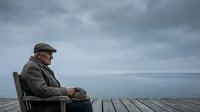Elderly man with gray hair and beard wearing brown cap and jacket sitting alone on wooden dock facing calm sea under cloudy sky, contemplative mood.