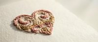Handmade rope heart decoration in pink and gold colors on neutral fabric background for craft projects and romantic designs.