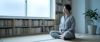 Young Asian woman practicing mindfulness meditation in modern home library with natural light, sitting cross-legged on floor for wellness and mental health.