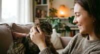 Young woman in cozy sweater gently petting tabby cat in warm living room with soft lighting. Pet care, companionship and relaxation at home concept.