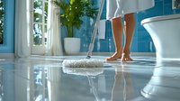 Woman mopping bathroom floor with white mop, bare feet visible against turquoise blue tiles, modern interior with potted palm plant and natural light.