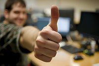 Close-up of hand showing thumbs up gesture against blurred office background, expressing positivity, approval and success in workplace environment.