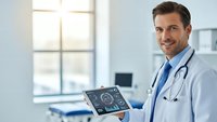 Confident male doctor holding digital tablet with medical data in modern hospital office, smiling at camera while wearing white coat and stethoscope.