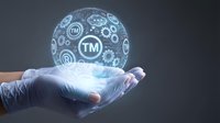 Gloved hand holding a glowing holographic sphere with trademark TM symbols and business icons, representing intellectual property protection.