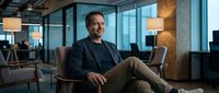 Confident middle-aged Caucasian businessman sitting relaxed in modern office chair, smiling warmly in contemporary corporate workspace with glass walls and city views.
