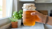 Woman holding glass jar of golden honey with twine bow in bright kitchen with potted plant and window light, suitable for food, cooking, and organic product marketing.
