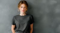 Young redhead woman with natural makeup wearing gray t-shirt against textured gray wall background, looking directly at camera with confident expression.