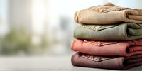 Neatly folded stack of cozy knitted sweaters in earthy tones of beige, olive green, and coral pink on blurred bright background. Perfect for seasonal fashion content.