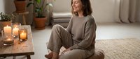 Young woman in comfortable beige loungewear sitting peacefully on floor with candles in cozy living room during relaxation time at home.