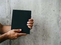 Male hands with medium skin tone holding a blank black book against a textured concrete wall background, suitable for publishing, education, and business concepts.