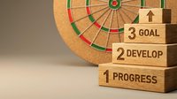 Wooden blocks stacked with progress, develop, and goal text next to dartboard, representing business strategy and achievement milestones for corporate planning.