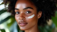 Young African woman with natural curly hair applying white sunscreen or skincare product to cheek, outdoor setting with blurred green foliage background.