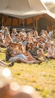 Young diverse crowd enjoying outdoor music festival sitting on grass in golden sunlight with stage tent in background during summer concert event.