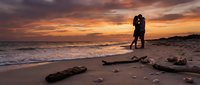 Romantic couple silhouette embracing on beach at sunset with driftwood and shells, golden hour sky reflecting on ocean waves for wedding anniversary concepts.
