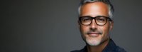 Confident mature businessman with gray hair and glasses wearing navy blue shirt against dark background, professional corporate headshot portrait.