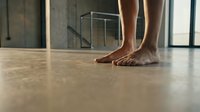 Barefoot woman standing on polished concrete floor in modern minimalist interior with glass railings and natural light streaming through windows.