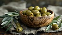 Fresh green olives in wooden olive bowl with olive branches on rustic linen cloth, moody natural lighting for Mediterranean food styling.