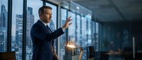 Professional businessman in dark suit pointing at city skyline through office windows at dusk, corporate leadership and urban business success concept.