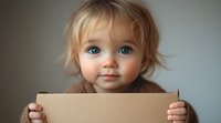 Adorable caucasian toddler with bright blue eyes and blonde hair holding cardboard sign, looking directly at camera with sweet expression against blurred background.