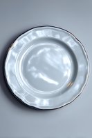 Empty white porcelain plate with gold trim and scalloped edges on gray background, photographed from above. Elegant dinnerware for fine dining and table setting.