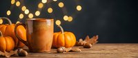 Warm copper mug with hot beverage surrounded by small pumpkins, nuts, and autumn leaves on wooden surface with golden bokeh lights, perfect for fall season marketing and holiday beverage promotions.