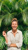 Young woman with reddish-brown hair in bun, fair complexion, smiling at camera against lush green palm foliage. Wearing light beige linen shirt. Tropical garden or botanical setting for lifestyle and nature themes.