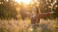 Young Caucasian woman with arms outstretched in a sunlit wildflower meadow, expressing joy, freedom, and harmony with nature at golden hour.