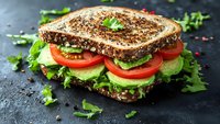 Fresh vegetable sandwich with whole grain bread, lettuce, tomatoes, cucumber slices and herbs on dark stone background, healthy lunch meal.